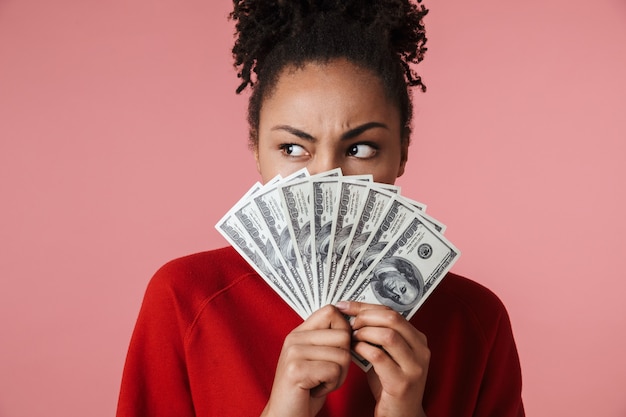 mooie geweldige jonge afrikaanse vrouw poseren geïsoleerd over roze muur met geld dat gezicht bedekt.