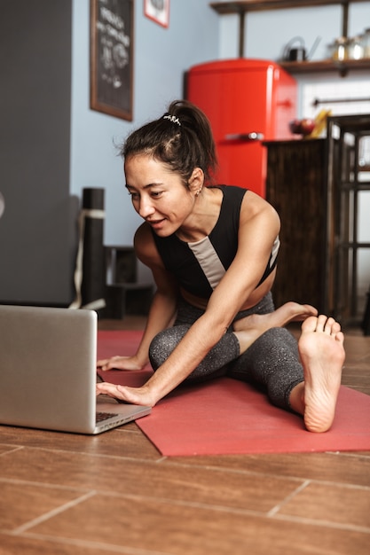 Mooie gezonde vrouw doen yoga oefeningen zittend op een fitness mat thuis, met behulp van laptop, stretching