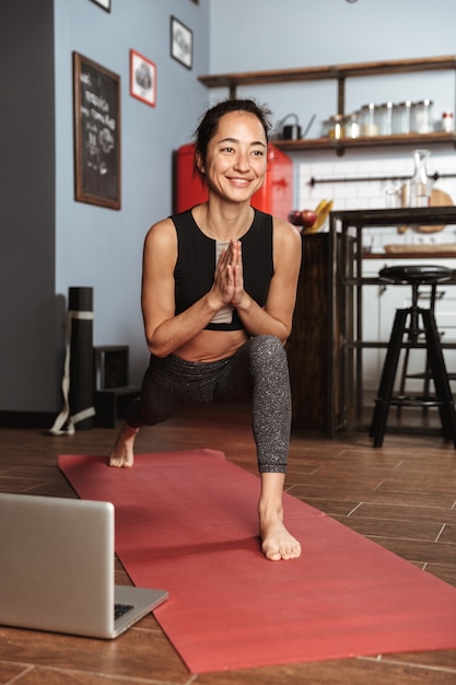 Mooie gezonde vrouw doen yoga oefeningen zittend op een fitness mat thuis, met behulp van laptop, stretching