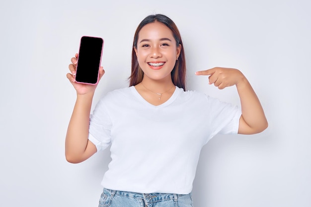 Mooie glimlachende Aziatische meisje dragen casual witte t-shirt wijzend een wijsvinger op mobiele telefoon met leeg scherm geïsoleerd op witte achtergrond People lifestyle concept