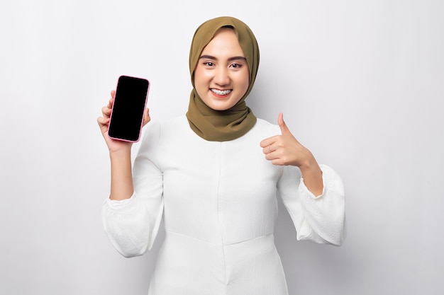 Mooie glimlachende Aziatische moslimvrouw die hijab draagt en mobiele telefoon vasthoudt met leeg leeg scherm met duim omhoog geïsoleerd op witte achtergrond studio portret Mensen religieuze levensstijl concept