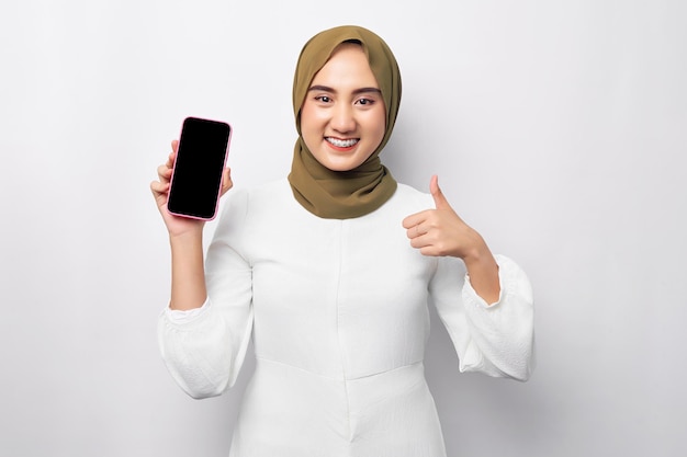 Mooie glimlachende aziatische moslimvrouw die hijab draagt en mobiele telefoon vasthoudt met leeg leeg scherm met duim omhoog geïsoleerd op witte achtergrond studio portret mensen religieuze levensstijl concept