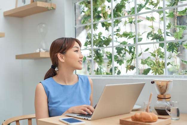 Mooie glimlachende Aziatische vrouw die achter het raam zit en op haar laptop leest.