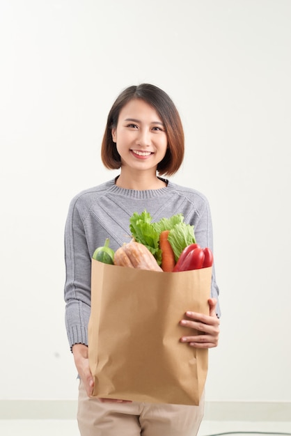 Mooie glimlachende Aziatische vrouw met papieren boodschappentas vol groenten en boodschappen, studio-opname geïsoleerd op een witte achtergrond