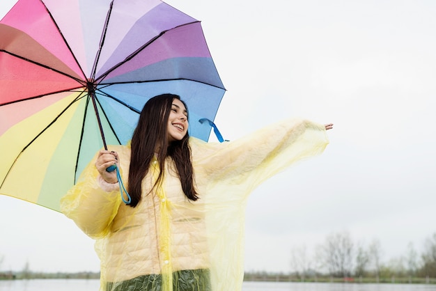 Mooie glimlachende brunette vrouw in gele regenjas met regenboogparaplu in de regen, regendruppels vangend met haar hand