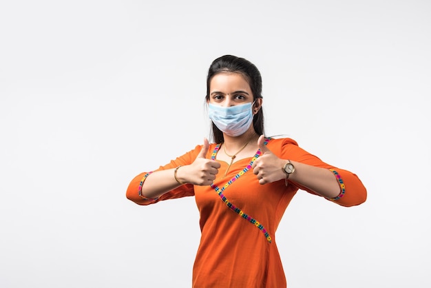 Mooie glimlachende Indiase jonge vrouw met beschermend gezichtsmasker, gelukkige duizendjarige vrouw in medische gezichtsbedekking van COVID-19 denken, gezondheidszorgconcept, geïsoleerd over witte muur