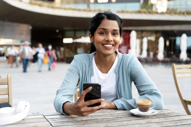 Mooie glimlachende Indiase vrouw met mobiele telefoon die koffie drinkt en naar de camera in café kijkt