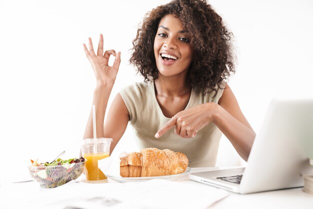 Mooie glimlachende jonge afrikaanse vrouw die aan het bureau zit geïsoleerd over een witte muur, luncht terwijl ze op een laptop werkt, met een goed gebaar