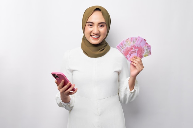 Mooie glimlachende jonge Arabische Aziatische moslimvrouw die hijab draagt die contant geld in rupiah-bankbiljetten en mobiele telefoon houdt geïsoleerd op witte achtergrond Mensen religieus levensstijlconcept