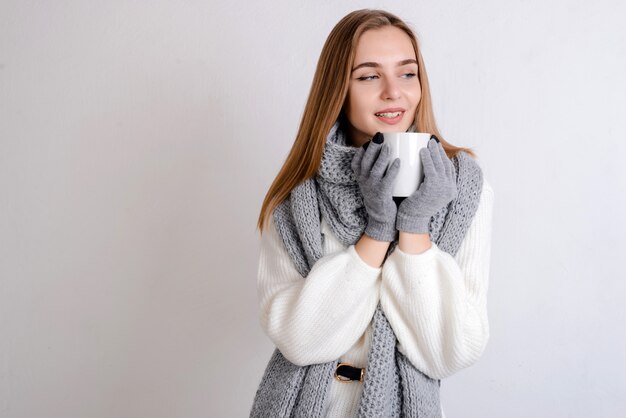 Mooie glimlachende jonge blondevrouw die in witte sweater, sjaal en handschoenen een witte kop van drank in haar handen houdt.