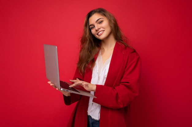 Mooie glimlachende jonge donkerbruine vrouw die netbookcomputer houdt die op toetsenbord typt die camera bekijkt die rood vest draagt