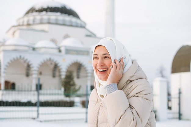 Mooie glimlachende jonge moslimvrouw met hoofddoek in lichte kleding die mobiel gebruikt tegen moskee