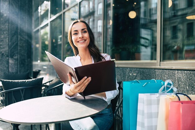 Mooie glimlachende jonge vrouw die in het straatcafé zit en het menu leest na het buiten winkelen