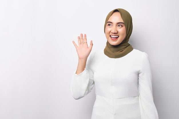 Mooie glimlachende vriendelijke jonge Arabische Aziatische moslimvrouw die hijab draagt zwaaiende hand die hallo zegt in lege ruimte geïsoleerd op witte achtergrond Mensen religieus levensstijlconcept