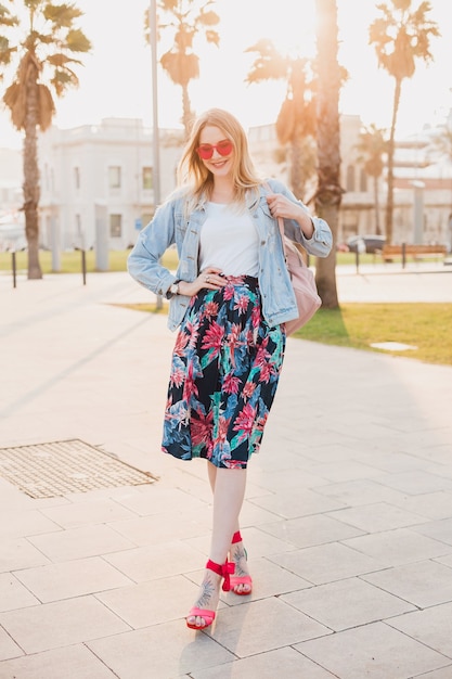 Mooie glimlachende vrouw die in de straat van de stad loopt in een stijlvolle bedrukte rok en een oversized denim jasje met een roze zonnebril