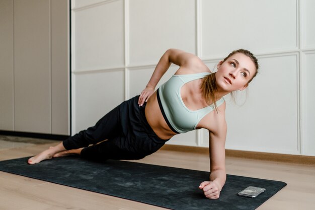 Mooie glimlachende vrouw die sportoefening met telefoon op yogamat thuis doet