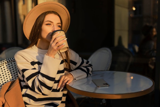 Mooie glimlachende vrouw in stijlvolle outfit zittend aan tafel in een hoed en trui romantische gelukkige stemming wachtend op een vriendje op een date in een café lente zomer modetrend koffie drinken