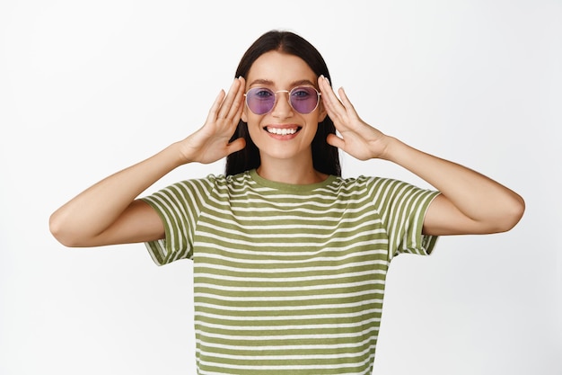 Mooie glimlachende vrouw zet een zonnebril op en ziet er gelukkig uit met zomerkleding op een witte achtergrond