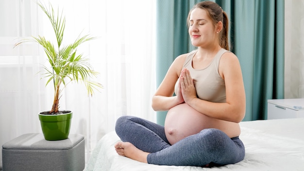 Mooie glimlachende zwangere vrouw die diep ademhaalt en mediteert in lotus asana op bed