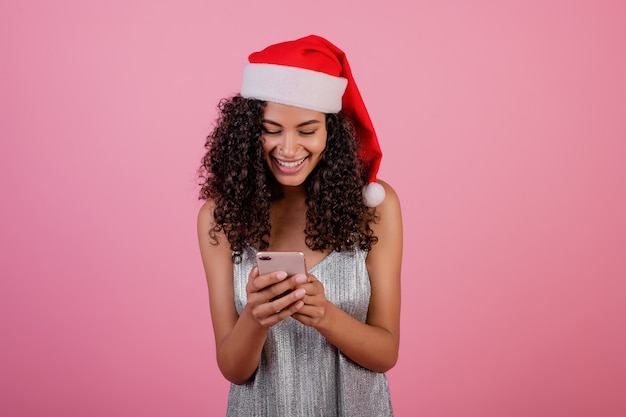 Mooie glimlachende zwarte met telefoon die kerstmishoed en kleding draagt die over roze wordt geïsoleerd