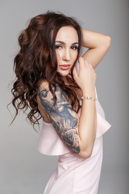 Mooie grappige vrouw met een kapsel met een tatoeage op haar arm in een roze avondjurk op een grijze achtergrond