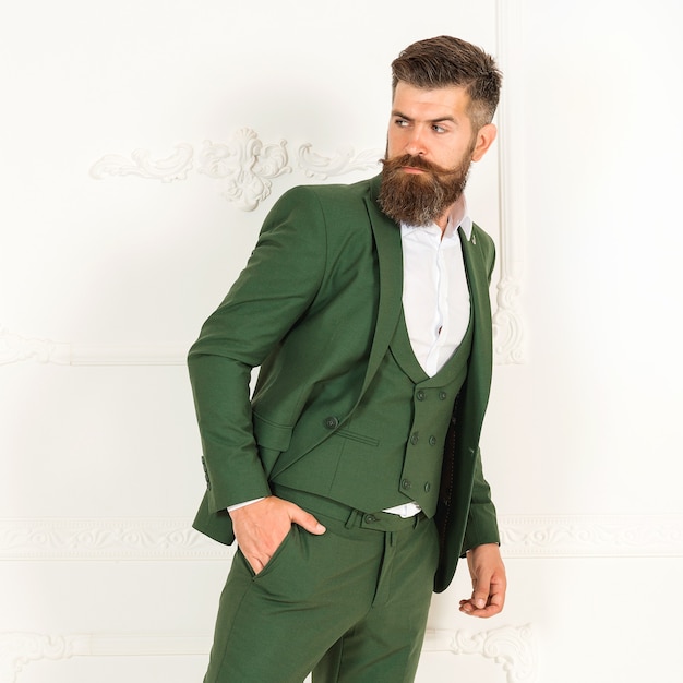 Mooie groene herenjas pak met shirt op brutale sexy bebaarde man. Mode portret van serieuze persoon met baard in groen elegant pak in witte kamer met klassiek interieur. Jonge mannequin