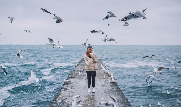 Mooie horizontale afbeelding van jonge vrouw voedt meeuwen op de zee Mooie vrouw draagt jas sjaal en kijken naar vliegende meeuwen door de zee in de lucht Mensen reizen milieu natuur concept