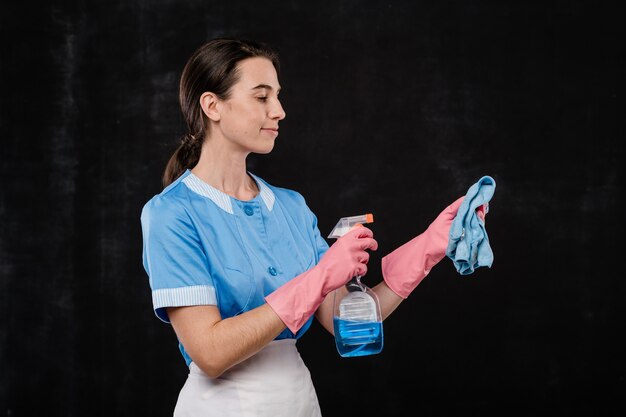 Mooie hotel meid in unifor en roze rubberen handschoenen afwasmiddel sproeien op stofdoek voor camera tegen zwarte achtergrond