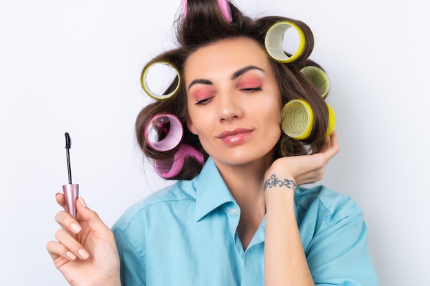 Mooie huisvrouw een jonge vrolijke vrouw met haarkrulspelden, felroze make-up en mascara bereidt zich voor op een avondje uit op een witte achtergrond