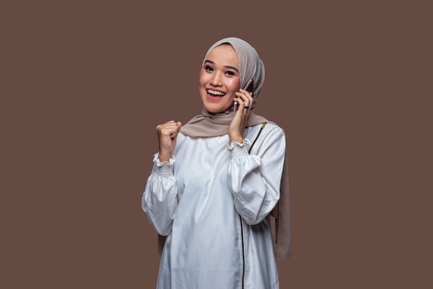 Mooie Indonesische vrouw die hijab draagt, krijgt het goede nieuws op haar mobiel met een glimlach en succesvolle uitdrukking geïsoleerd op effen achtergrond