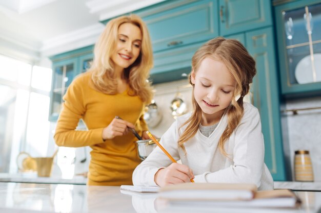 Mooie inhoud blonde meisje glimlachend en schrijven in haar notitieboekje terwijl haar moeder achter haar met een steelpan