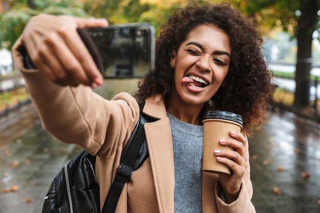 Mooie jonge afrikaanse vrouw dragen jas buiten wandelen in het park, met rugzak, een selfie te nemen