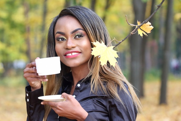 Mooie jonge Afrikaanse vrouw koffie drinken uit een witte kop op de ruimte van de herfstbladeren in het Park