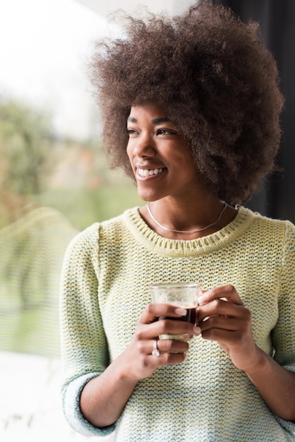 Mooie jonge Afro-Amerikaanse vrouw die koffie drinkt en door een raam kijkt in haar luxe huis