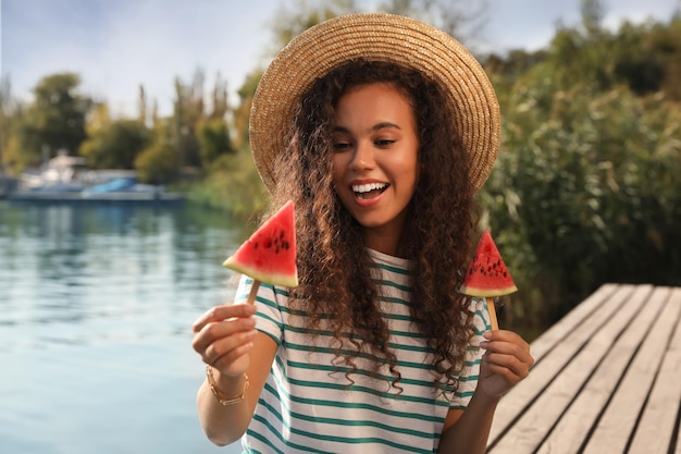 Mooie jonge Afro-Amerikaanse vrouw met stukjes watermeloen op houten pier in de buurt van rivier