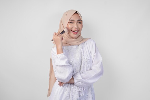 Foto mooie jonge aziatische moslimvrouw met een witte jurk en hijab die make-up aanbrengt met behulp van een penseel mode en cosmetica schoonheidsconcept