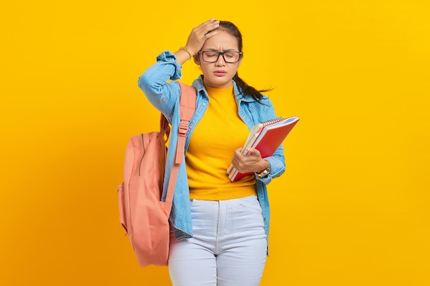 Mooie jonge Aziatische student in denim outfit met rugzak notebook houden en op zoek duizelig met hoofdpijn op gele achtergrond Onderwijs in middelbare school universiteit college concept