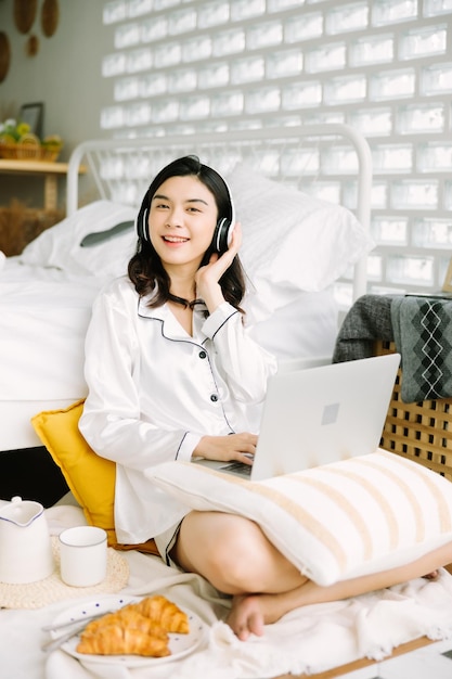 Mooie jonge Aziatische vrouw die naast bed zit met laptop en ontbijt in de ochtend thuis