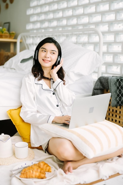 Mooie jonge Aziatische vrouw die naast bed zit met laptop en ontbijt in de ochtend thuis