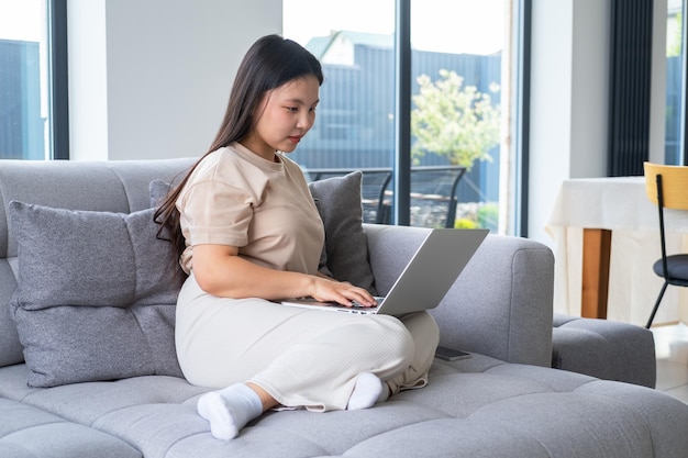 mooie jonge aziatische vrouw in casual, gezellige kleding met behulp van laptop zittend op de bank in modern appartement