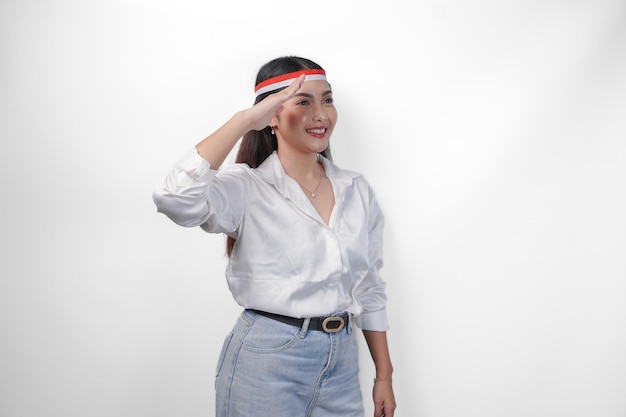 Mooie jonge Aziatische vrouw in een casual outfit en met een vlag op haar hoofdband die een groet gebaar maakt met een kopie ruimte geïsoleerd op een witte achtergrond Indonesiese onafhankelijkheidsdag concept