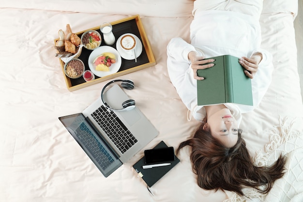 Mooie jonge Aziatische vrouw liggend op bed naast laptop en ontbijtblad en denken na het lezen van een tot nadenken stemmend artikel in boek