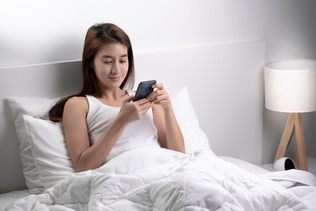 Mooie jonge Aziatische vrouw ontspant in de slaapkamer en wordt 's ochtends wakker met sms'jes op de telefoon