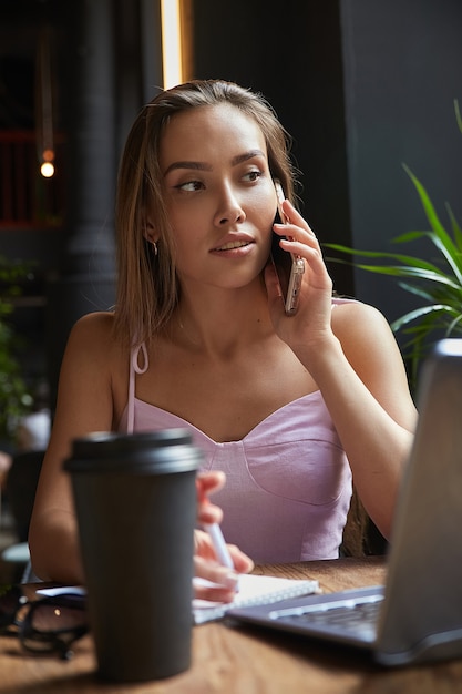 Mooie jonge Aziatische vrouw zit met laptop in café bellen met smartphone