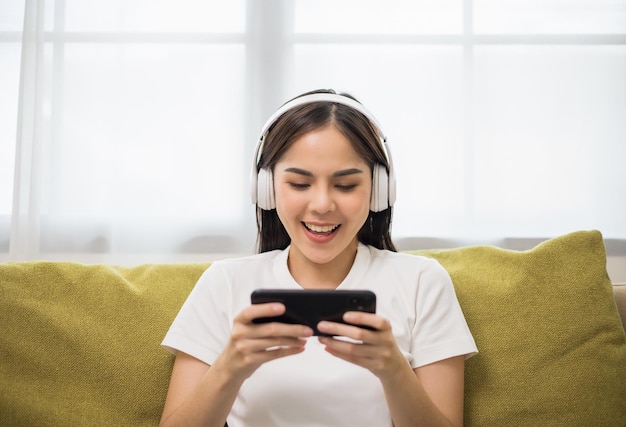 Mooie jonge aziatische vrouwen spelen mobiel spel en zetten een draadloze hoofdtelefoon op die op de bank in de woonkamer zit. Spel spelen op smartphone winnend overwinningsmoment. Heel veel plezier en leuke relaxtijd