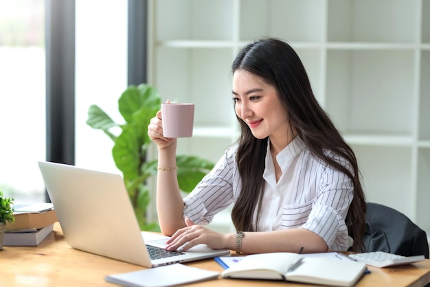 Mooie jonge Aziatische zakenvrouw die een kopje koffie drinkt die op laptop op kantoor werkt.