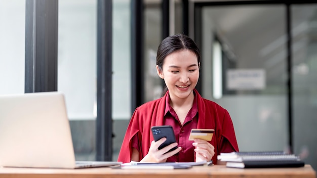 Mooie jonge Aziatische zakenvrouw in rood shirt glimlachend gelukkig winkelen online met smartphone en creditcard op kantoor.