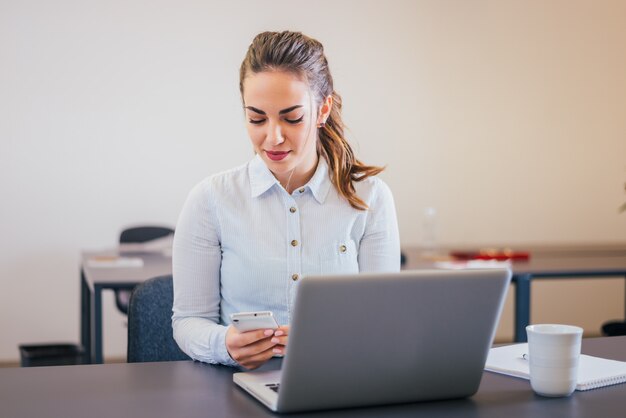 Mooie jonge bedrijfsvrouw die de telefoon bekijkt terwijl het zitten voor laptop