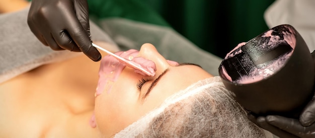 Mooie jonge blanke vrouw die een alginisch masker op het gezicht krijgt in de schoonheidssalon Gezichtsbehandeling van de huid
