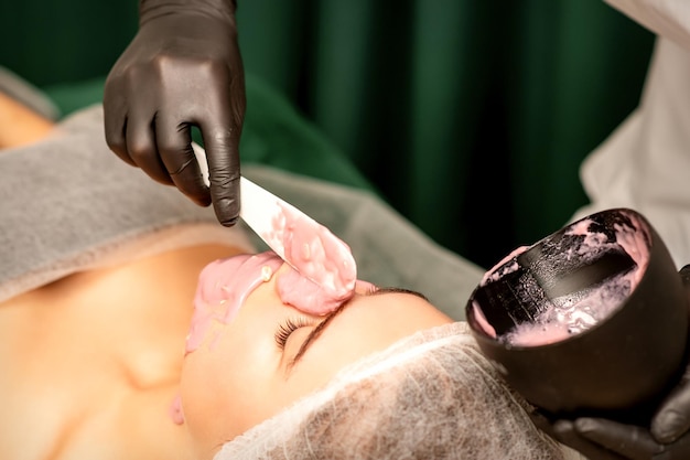 Mooie jonge blanke vrouw die een alginisch masker op het gezicht krijgt in de schoonheidssalon Gezichtshuidbehandeling